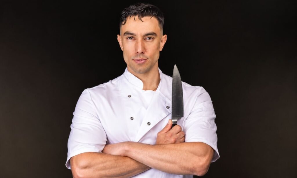 Professional chef wearing a white chef’s jacket, holding a kitchen knife against a dark studio background with dramatic lighting.