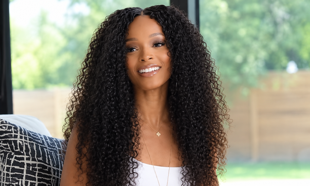 Loreal Sarkisian with long curly hair wearing a white top and gold necklace, seated indoors with a bright garden view in the background.