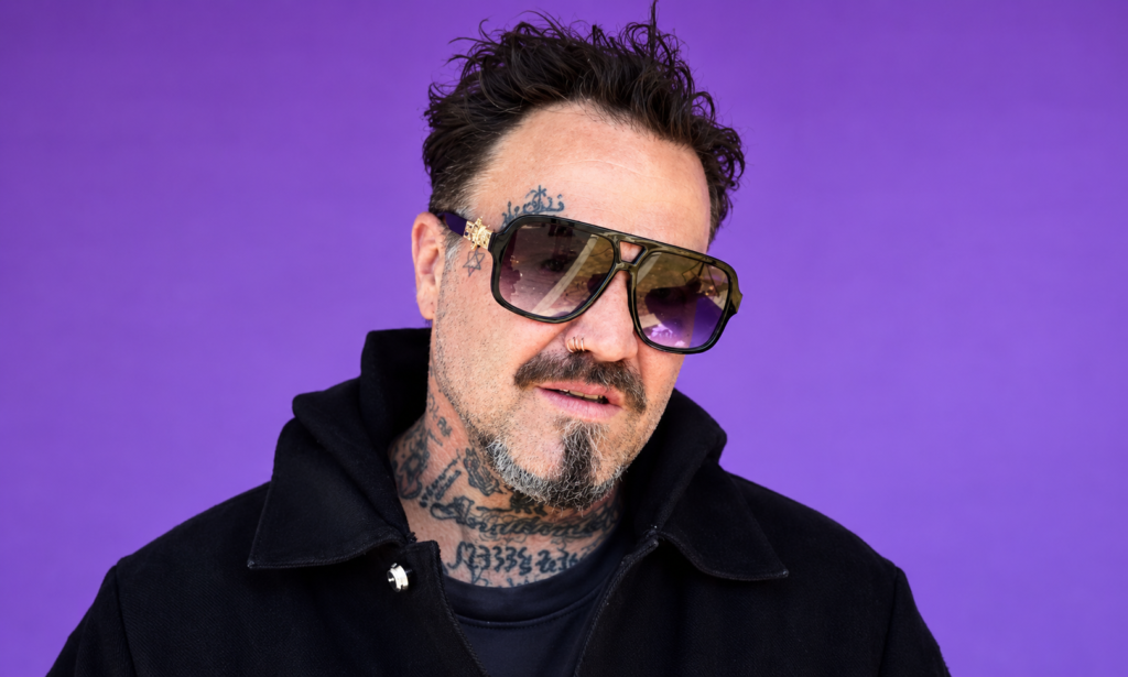 Bam Margera wearing sunglasses and a black jacket, shown in a close-up portrait against a purple background.