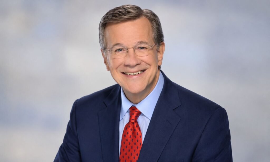 David Asman wearing a navy blue suit, light blue shirt, and red patterned tie, smiling in a professional portrait against a light gray background.
