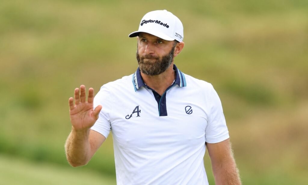 Dustin Johnson wearing a white TaylorMade golf cap and polo shirt, raising his hand on a golf course during a professional golf event.