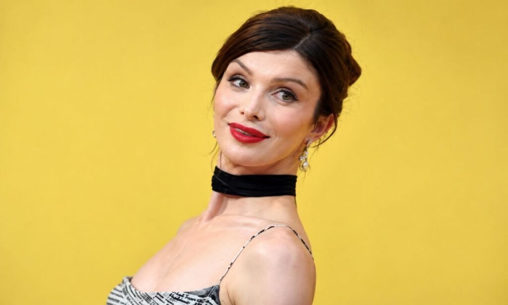 Dylan Mulvaney posing in an elegant black choker and patterned dress against a clean yellow background at a red-carpet event.