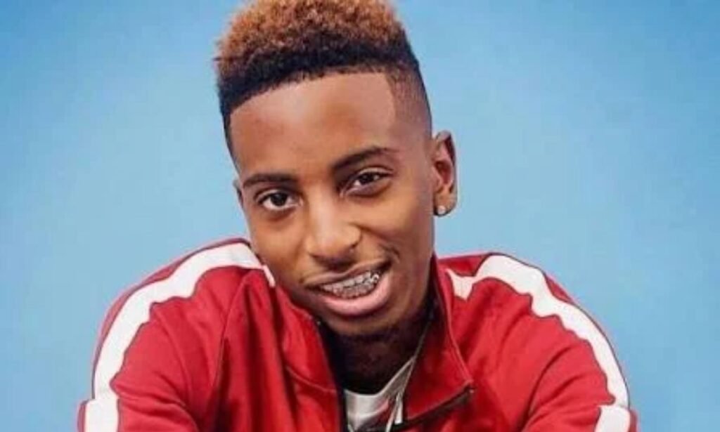 FunnyMike wearing a red Nike tracksuit and silver medal, smiling in a blue studio portrait.