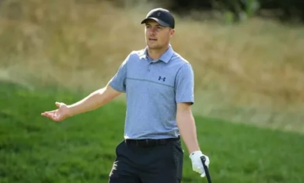 Jordan Spieth wearing a blue polo shirt and black cap on a golf course during a professional golf event.