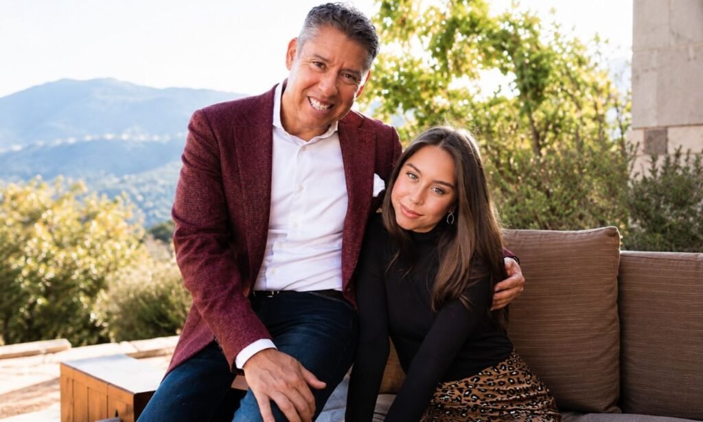 Luis Strohmeier posing with a young woman outdoors, wearing a burgundy blazer and white shirt in a sunlit setting with a scenic natural background.