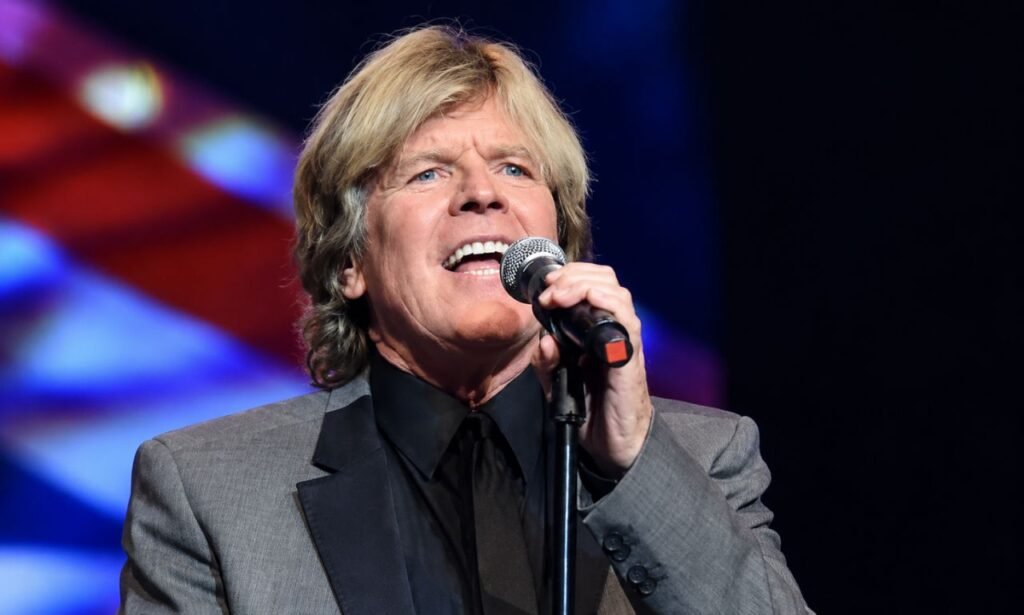Peter Noone performing live on stage in a gray suit, singing into a microphone under colorful concert lights.