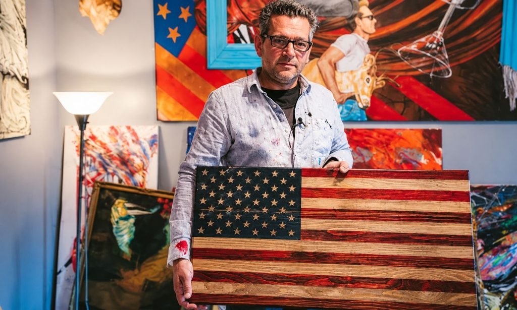 Scott LoBaido holding a rustic American flag artwork inside his colorful art studio, surrounded by paintings and creative artwork.