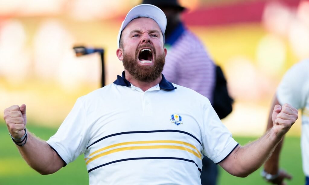 Shane Lowry celebrating a golf victory with clenched fists, wearing a white cap and Ryder Cup polo shirt on the golf course.
